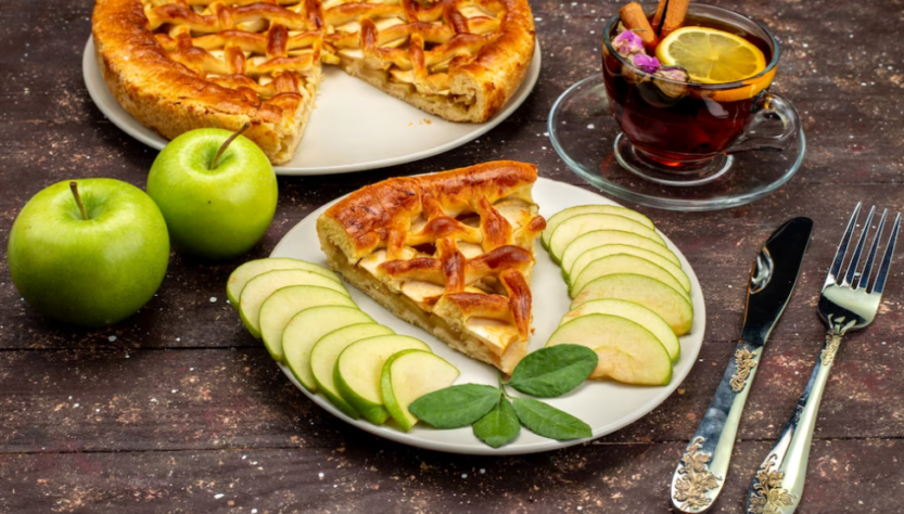 apple cake, a cup of tea, fresh green apples, fork, and spoon on the wooden desk