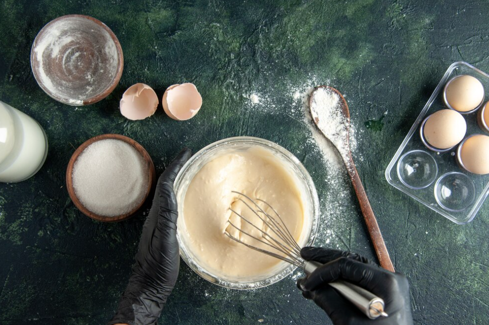 hand in black gloves making a dough with dough whisk, spoon with flour, eggs, and sugar on the green table