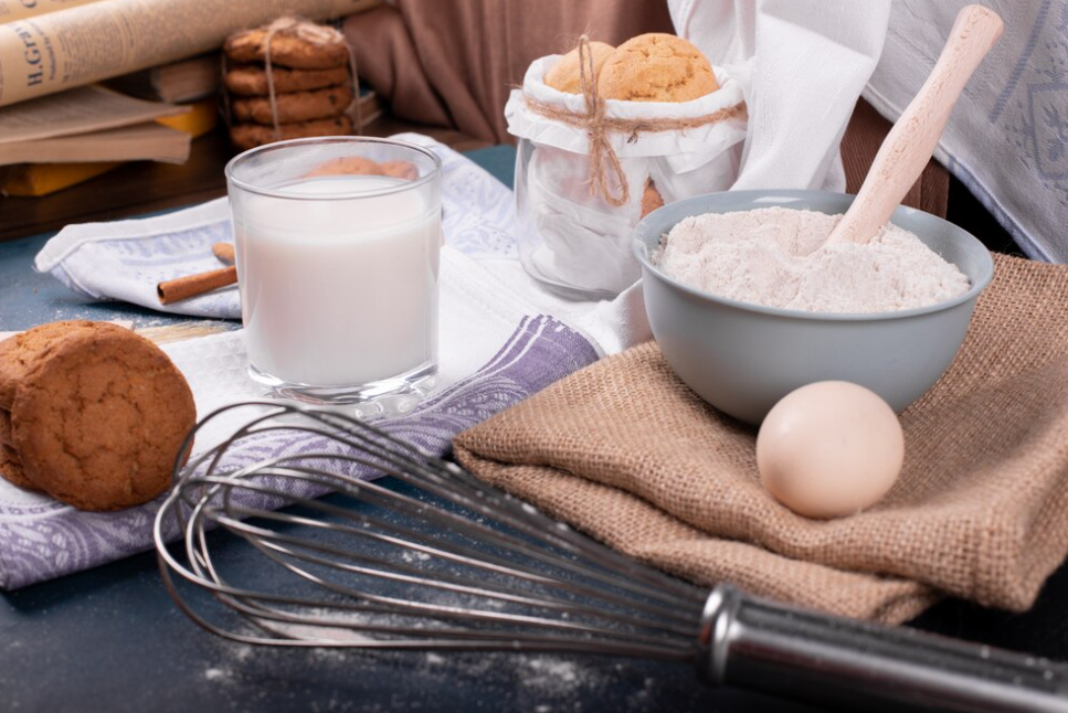 cup of milk, flour in a bowl, cookies,dough whisk and egg on the table